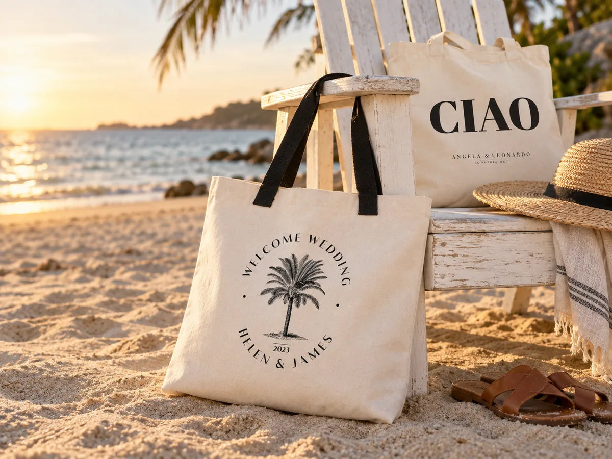 Strand zur goldenen Stunde: große Canvas-Tasche „Welcome Wedding / Helen & James“ mit Palmen-Emblem und schwarzen Henkeln lehnt an einem weißen Adirondack-Stuhl, dahinter eine „CIAO“-Tasche, ein Strohhut und Ledersandalen